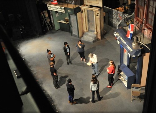 A theater rehearsal scene with seven people gathered around a director on a stage set resembling a city street.