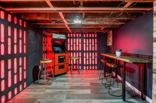 A retro arcade setup featuring a Pac-Man machine, bar stools, and a wooden table in a dimly lit room with red accents.
