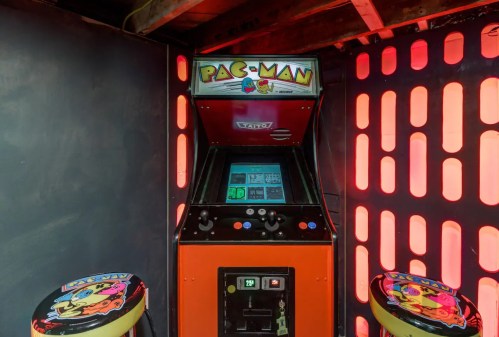 A classic Pac-Man arcade machine with colorful graphics, flanked by two stools, set against a lit backdrop.