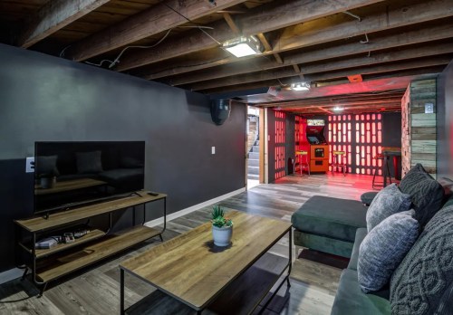 A modern basement living area with a TV, coffee table, and a cozy couch, featuring exposed beams and colorful lighting.