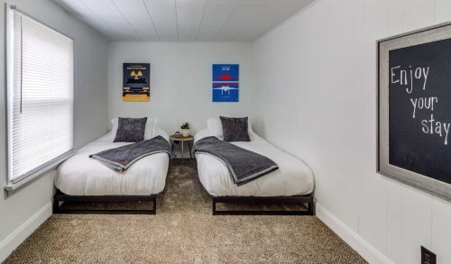 A cozy room with two twin beds, gray blankets, and wall art, featuring a chalkboard that says "Enjoy your stay."