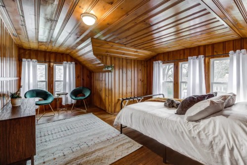 Cozy bedroom with wooden walls, a bed, two chairs, and large windows letting in natural light.