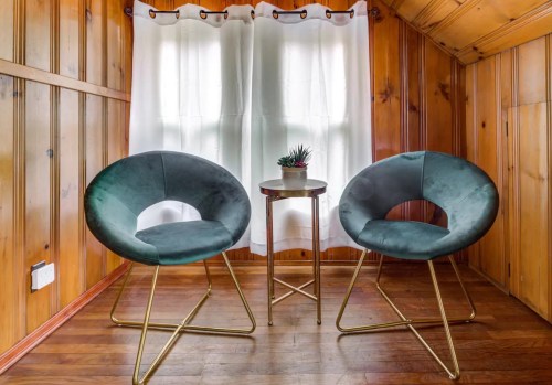 Two green chairs with gold legs and a small table with a potted plant, set against a wooden wall and white curtains.