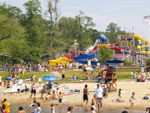 A busy beach scene with families enjoying the sun, water slides in the background, and people relaxing on the sand.