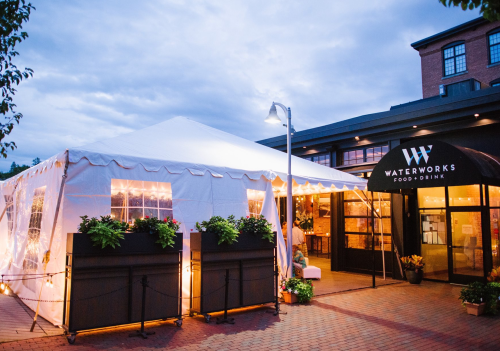 A tented outdoor dining area with string lights, next to a modern restaurant entrance. Evening sky in the background.