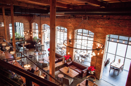 A spacious restaurant with brick walls, large windows, wooden beams, and cozy seating arrangements.