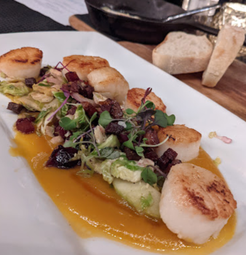 Seared scallops on a bed of greens with bacon, microgreens, and a drizzle of sauce, served with slices of bread.