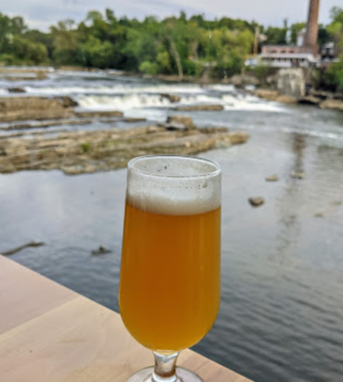 A glass of beer sits on a table with a scenic view of a river and waterfall in the background.