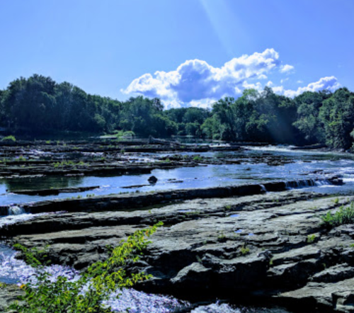 A serene river scene with rocky banks, gentle waterfalls, and lush greenery under a bright blue sky with fluffy clouds.