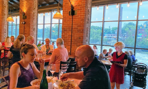 A cozy restaurant scene with diners enjoying meals by large windows overlooking a river. Warm lighting and brick walls.