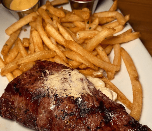 A juicy steak topped with butter, served alongside crispy French fries and dipping sauces.