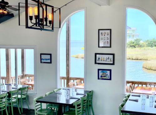 Bright restaurant interior with green chairs, large windows overlooking a scenic water view, and framed artwork on the walls.