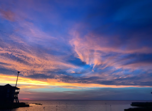 A vibrant sunset over the water, with colorful clouds and a silhouette of a building on the shore.
