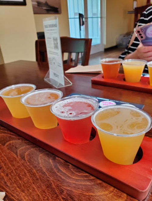 A wooden flight board with four small cups of different colored drinks, set on a table in a casual dining space.