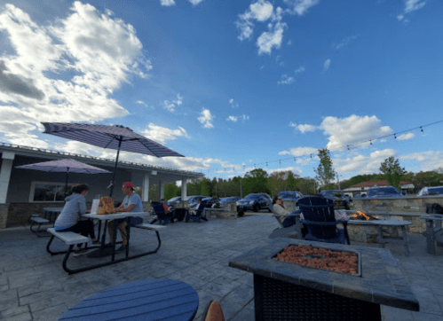 Outdoor seating area with umbrellas, people dining, and a fire pit under a blue sky with scattered clouds.