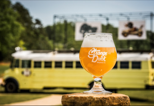 A glass of beer sits on a stone, with a yellow bus and banners in the background on a sunny day.