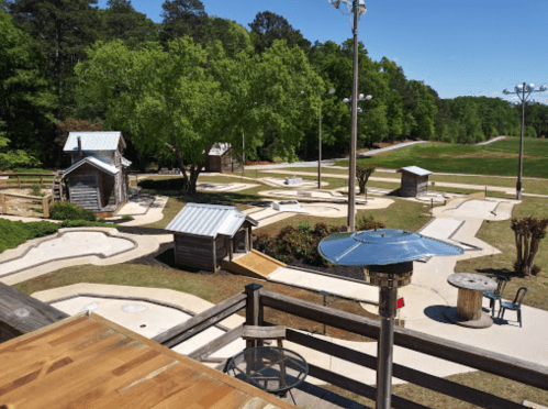 Aerial view of a mini-golf course with wooden structures, surrounded by trees and a clear blue sky.