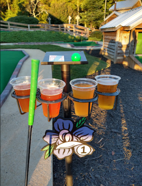 A mini-golf setup with a scorecard, a green golf ball, and four cups of different colored drinks on a holder.