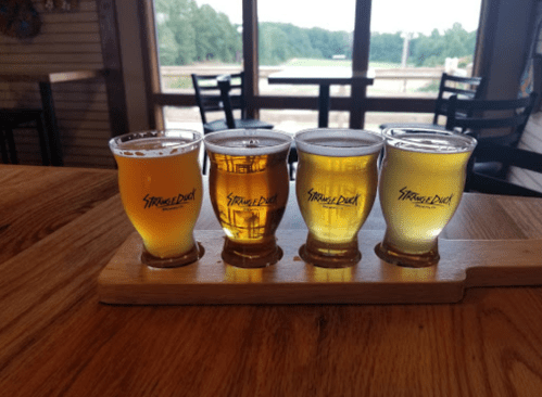 Four small glasses of beer on a wooden flight paddle, with a scenic view in the background.