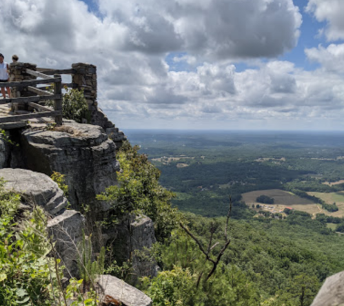 A scenic view from a rocky overlook, featuring lush green hills and a cloudy sky in the background.