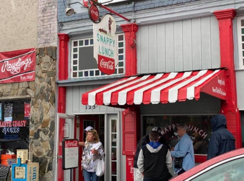 A red and white striped awning marks the entrance of Snappy Lunch, with people waiting outside in rainy weather.