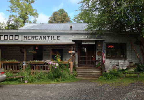 A rustic mercantile store with a bakery sign, surrounded by greenery and flowers, featuring an "Open" neon sign.