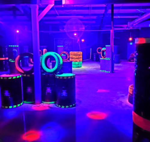 A dimly lit indoor laser tag arena with colorful neon lights, barrels, and tires arranged as obstacles.