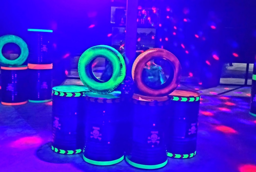 Colorful barrels with glowing rims and inflatable rings, illuminated by vibrant lights in a dark setting.