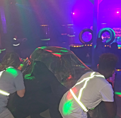 Two players in a laser tag arena crouch behind a camouflage barrier, with colorful lights illuminating the scene.