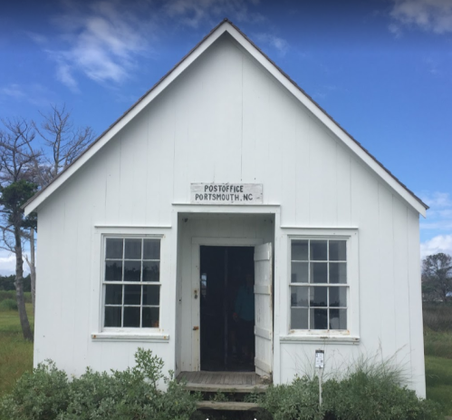 A small white building with a sign reading "Post Office Portsmouth, NC," surrounded by greenery and blue skies.