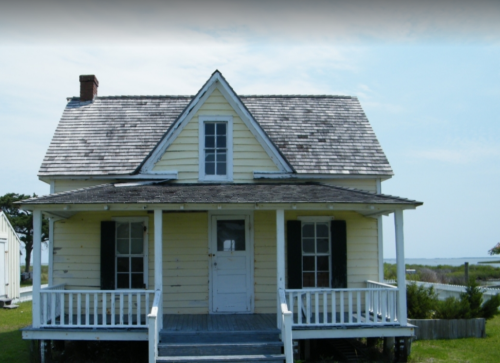 A yellow, two-story house with a gabled roof and a front porch, set against a coastal backdrop.