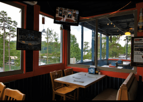 Cozy restaurant interior with wooden tables, large windows, and a view of a lake surrounded by trees.