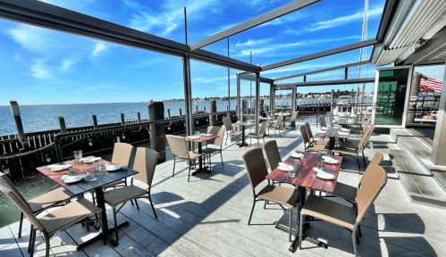 Outdoor dining area with tables set for guests, overlooking a calm waterfront under a clear blue sky.
