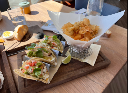 Three tacos with toppings on a wooden platter, accompanied by a basket of crispy chips and a lime wedge.