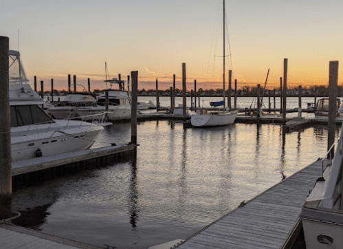 Sunset over a marina with boats docked and reflections on the water, surrounded by wooden posts.