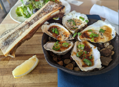 A cast iron dish with baked oysters topped with green onions, served with a side of bone marrow and a lemon wedge.