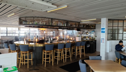 A modern bar area with high stools, a glass display of bottles, and a cozy dining space in a bright restaurant.