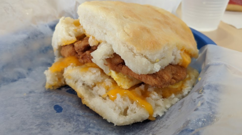 A close-up of a biscuit sandwich filled with fried chicken, melted cheese, and an egg, served on a blue plate.