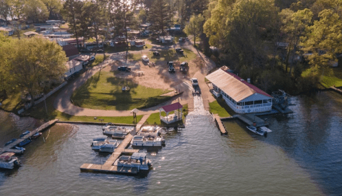 Aerial view of a lakeside area with boats, a dock, and a restaurant surrounded by trees and grassy spaces.