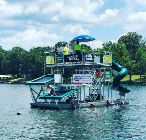 A floating water park with slides and sunshades on a lake, surrounded by trees and blue skies.