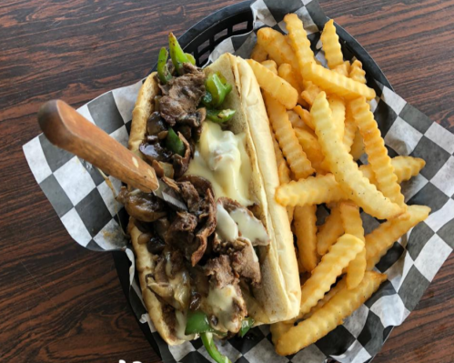 A loaded cheesesteak sandwich with peppers and onions, served with crinkle-cut fries in a checkered basket.