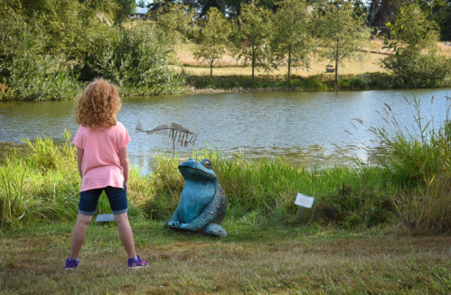 A child with curly hair stands by a pond, gazing at a large frog sculpture amidst greenery.
