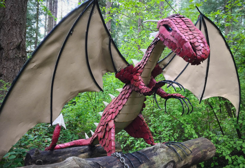 A red dragon sculpture with large wings stands on a log in a lush green forest.