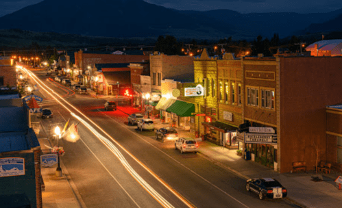 A charming small town street at night, with glowing storefronts and light trails from passing cars.