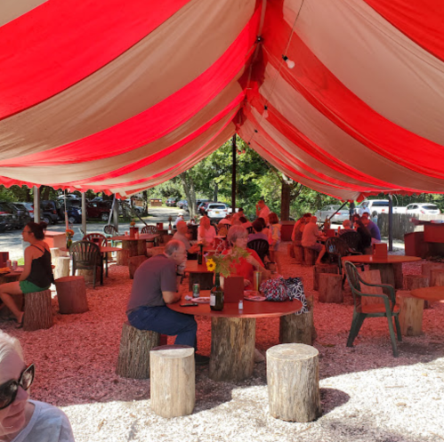 Outdoor dining area with red and white striped tent, wooden tables, and patrons enjoying their meals.