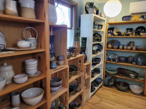 A cozy pottery studio with wooden shelves displaying various ceramic pieces, including bowls, vases, and mugs.