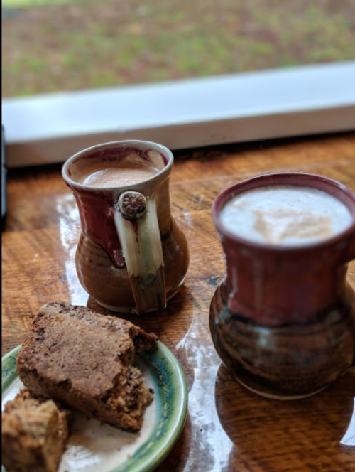 Two mugs of coffee beside a plate of brown baked goods on a wooden table, with a window in the background.