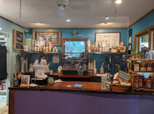 A cozy café interior with a counter, coffee machine, shelves of drinks, and a barista preparing orders.