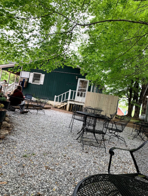 A gravel patio with empty metal tables and chairs, surrounded by greenery and a blue building in the background.