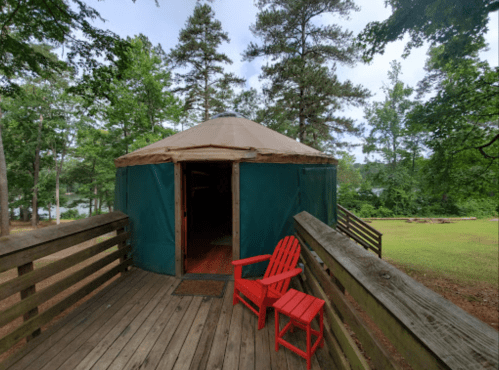 A yurt with a green exterior and wooden deck, featuring two red chairs, surrounded by trees and grass.